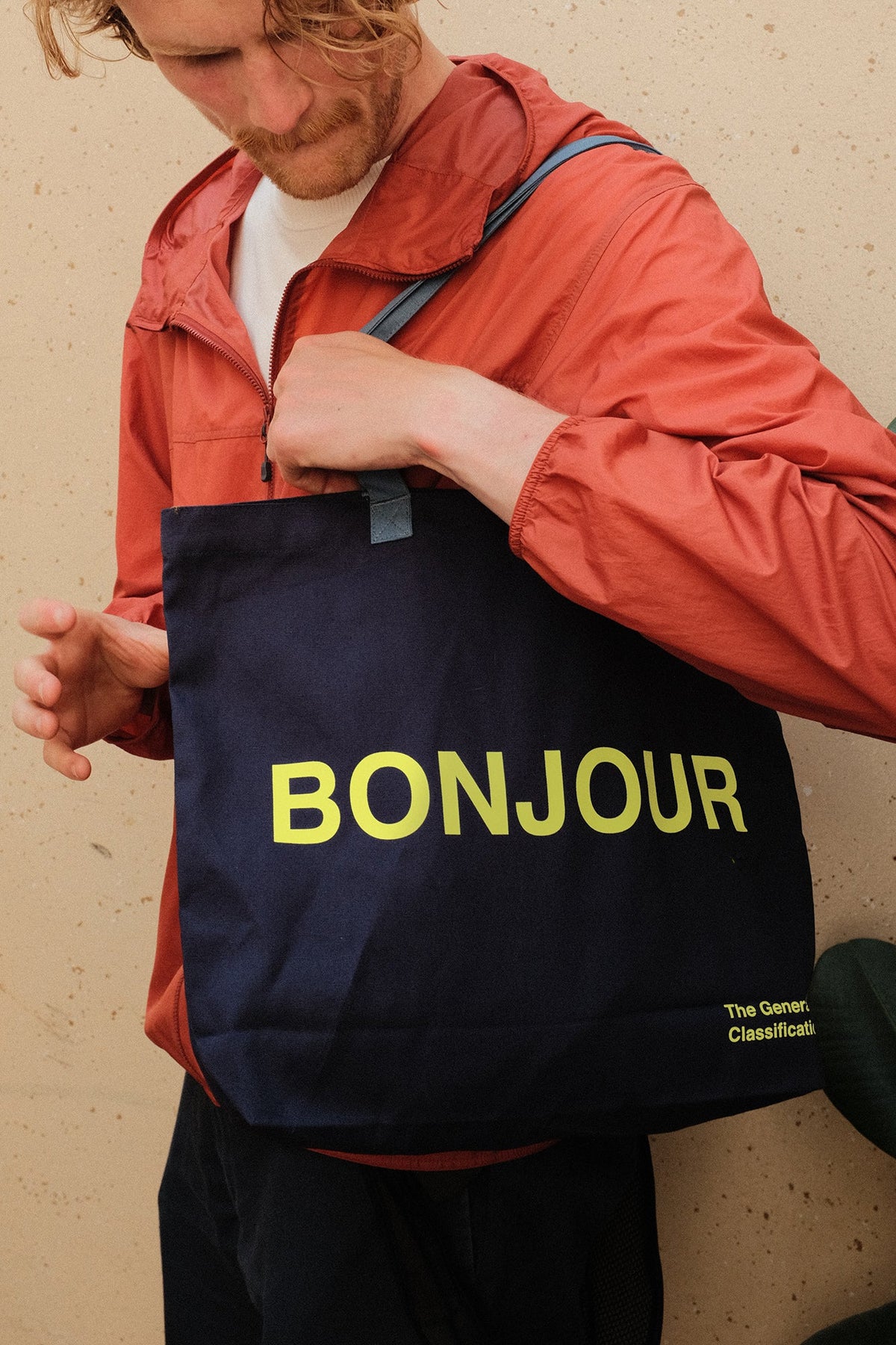 A man wearing a rust-orange windbreaker jacket carries a navy blue tote bag with 'BONJOUR' printed in yellow across the front. The brand 'The general classification' is visible in small text in the bottom right corner of the bag.
