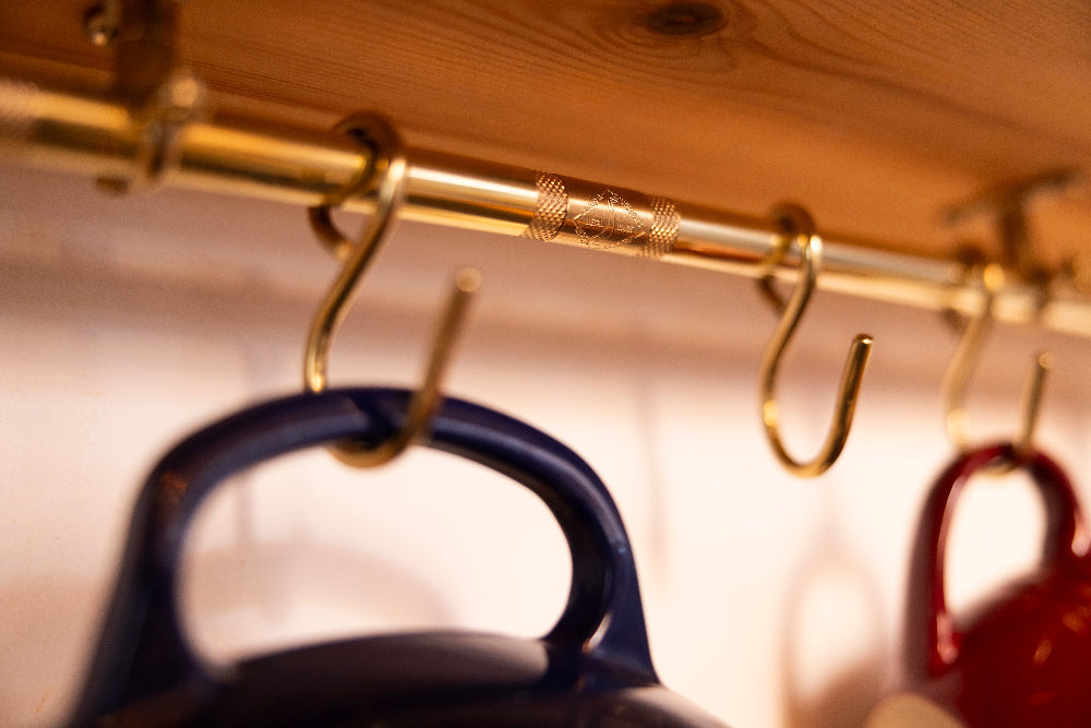 A brass hanging rod with multiple hooks mounted on a wooden shelf, with kettle or pot handles hanging below in blue and red.