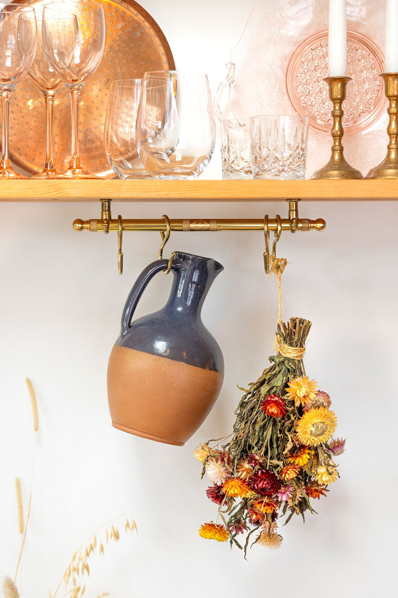 A rustic ceramic pitcher with a dark blue glazed upper half and a terracotta lower half hangs from a brass rack on a white wall. To the right of the rack, a small bundle of dried flowers hangs from the same brass hook.