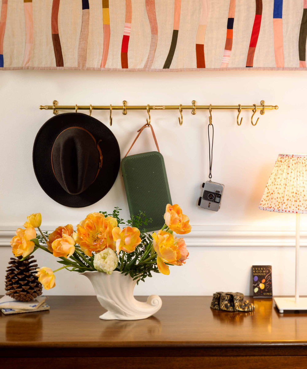 A brass wall-mounted coat rack with multiple hooks styled in a home interior. A black wide-brim hat hangs on the left, a green rectangular bag hangs in the center, and a small camera hangs on the right.