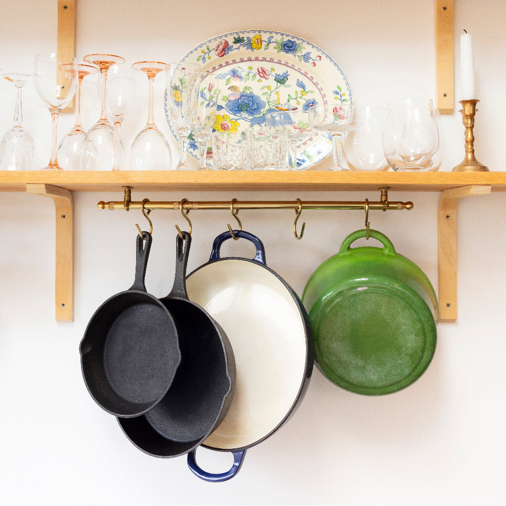 A set of cookware, including two black cast-iron skillets, a cream-colored Dutch oven, and a green pot, hangs from brass hooks on a wooden shelf below decorative glassware and a floral serving platter.