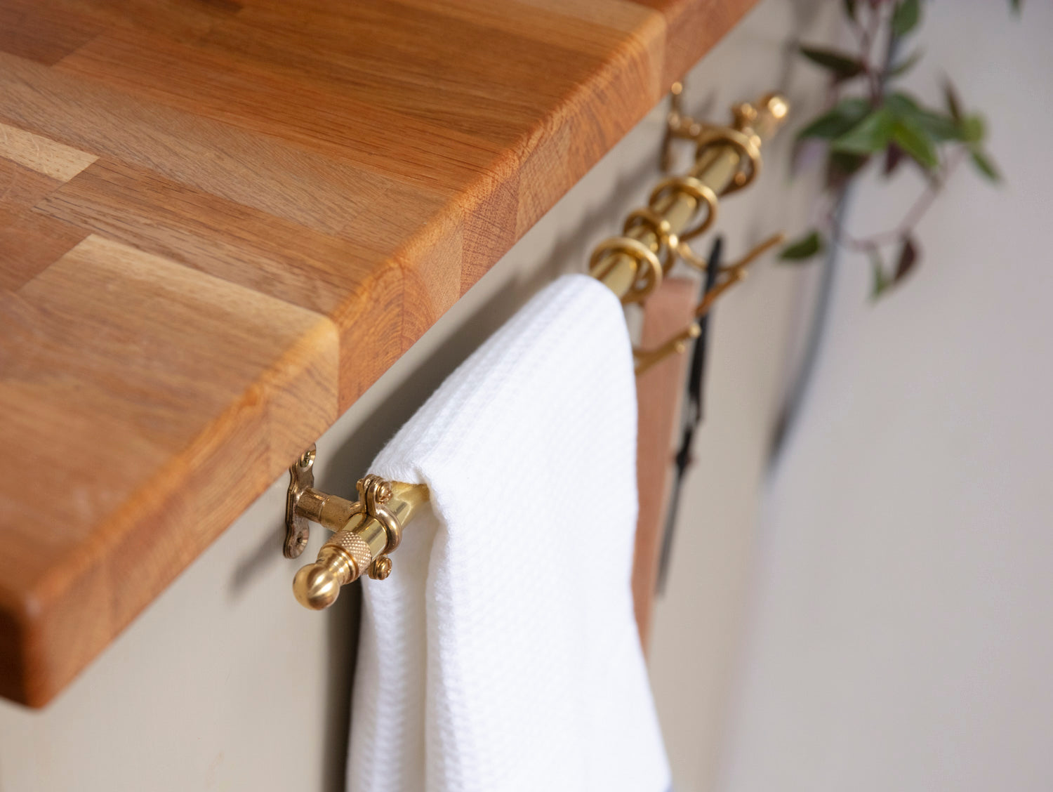 A close-up of a brass towel bar mounted on a light wall, holding a white textured towel. A wooden countertop is visible in the foreground, with a blurred green plant in the background.