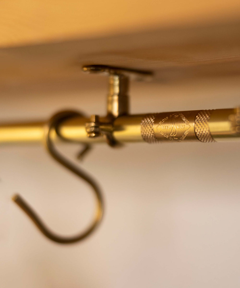 Close-up of a brass hanging rod with a decorative knurled section and a metal hook, shown against a warm beige background.