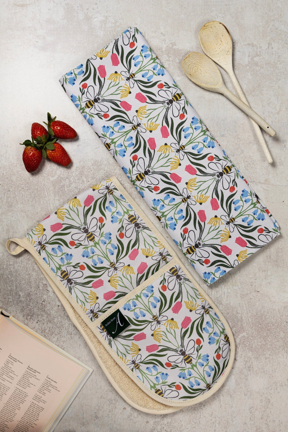 Floral-patterned oven mitt and tea towel on a textured surface with strawberries and wooden spoons.