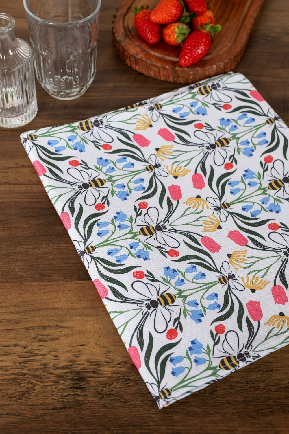 Floral and bee-patterned cloth on a wooden surface with strawberries and glasses in the background