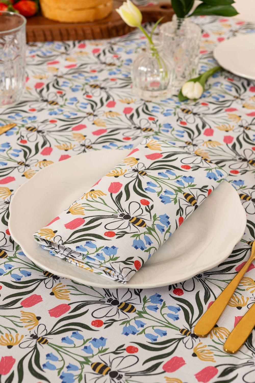 Table setting with floral tablecloth, plates, and folded napkin featuring bees and flowers.