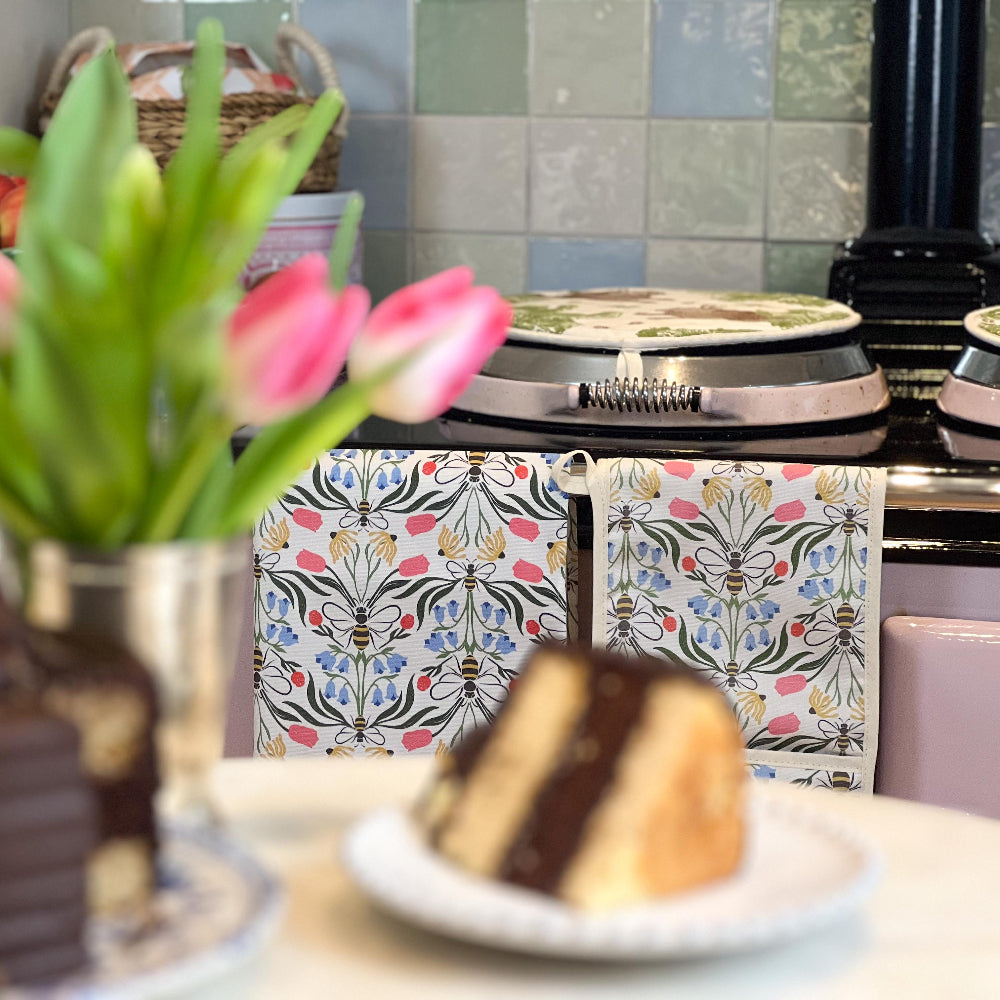 Decorative kitchen scene with a floral-patterned oven mitt and tea towel, tulips, and a cake.