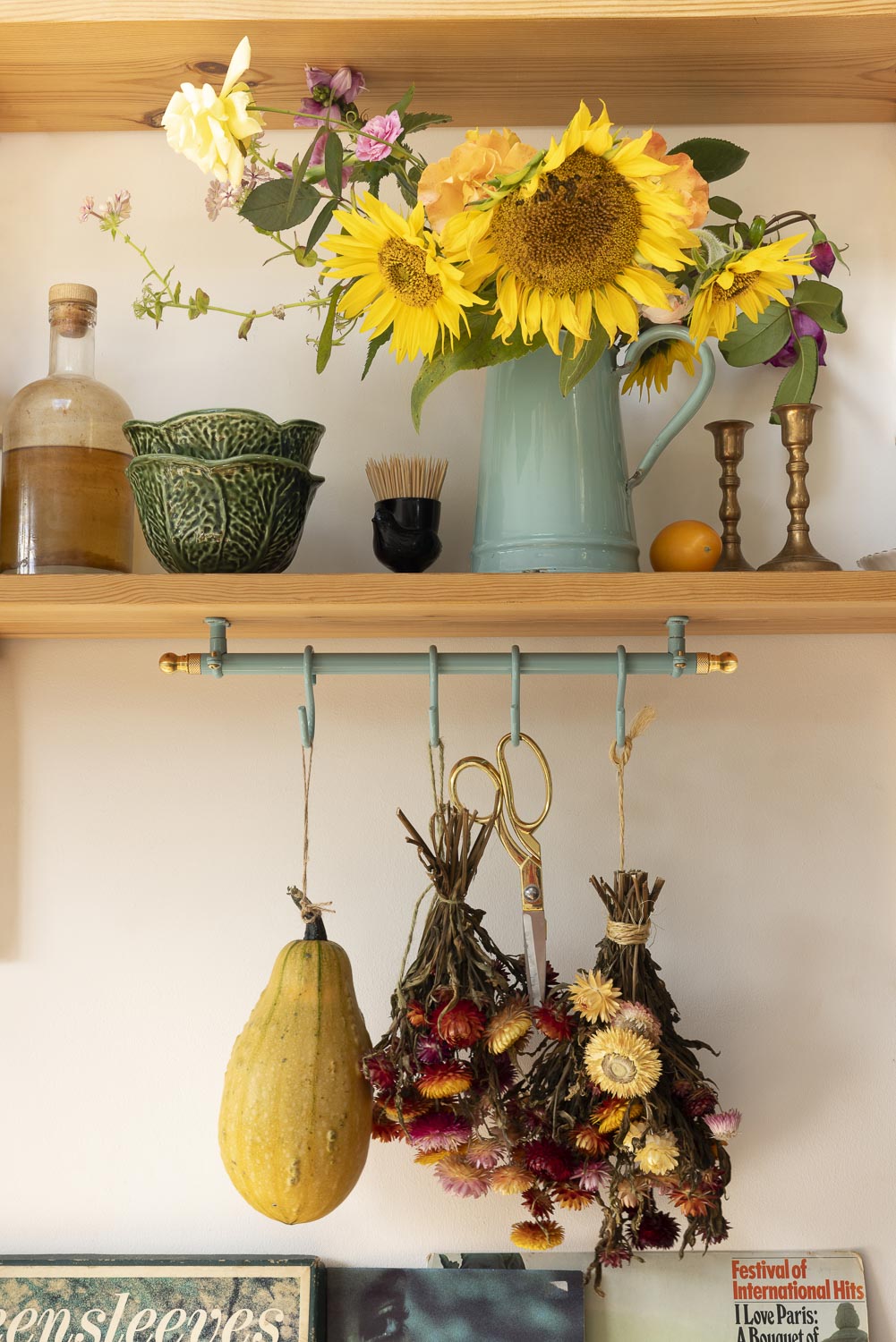 A styled interior shelf scene with wooden shelves, a mint green pitcher filled with bright yellow sunflowers, decorative bottles and vessels, and dried flower bundles hanging from a teal rack on the wall.