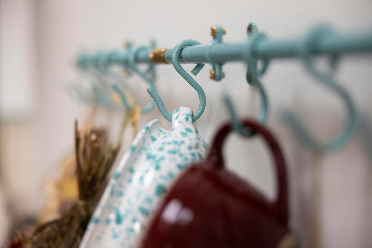 A close-up of a light blue metal hanging rod with multiple S-hooks, with a white patterned cloth and a dark red mug hanging from the hooks.