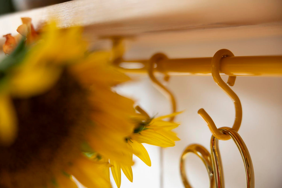 Close-up of a yellow metal rack rod with decorative S-shaped hooks, partially obscured by blurred yellow flowers in the foreground.