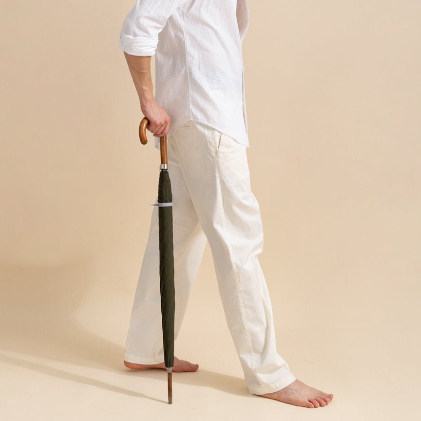 Person wearing white pants and a white shirt holding a racing green umbrella on a beige background