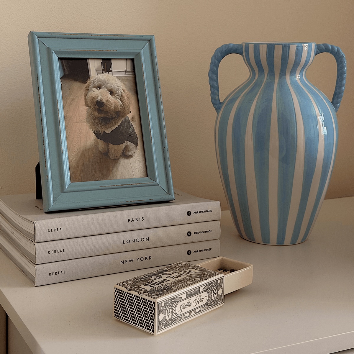 Gallia Rae Buon Natale matchbox styled on a shelf beside stacked books, a blue picture frame and striped ceramic vase