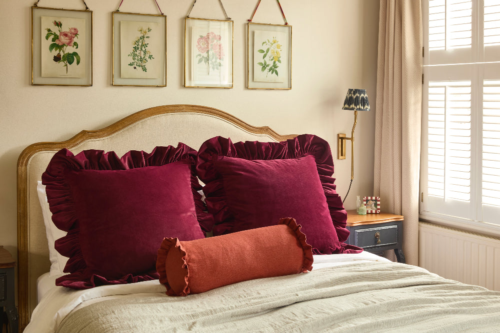 Bedroom with burgundy ruffled pillows on a bed, framed botanical art on the wall, and a lamp.
