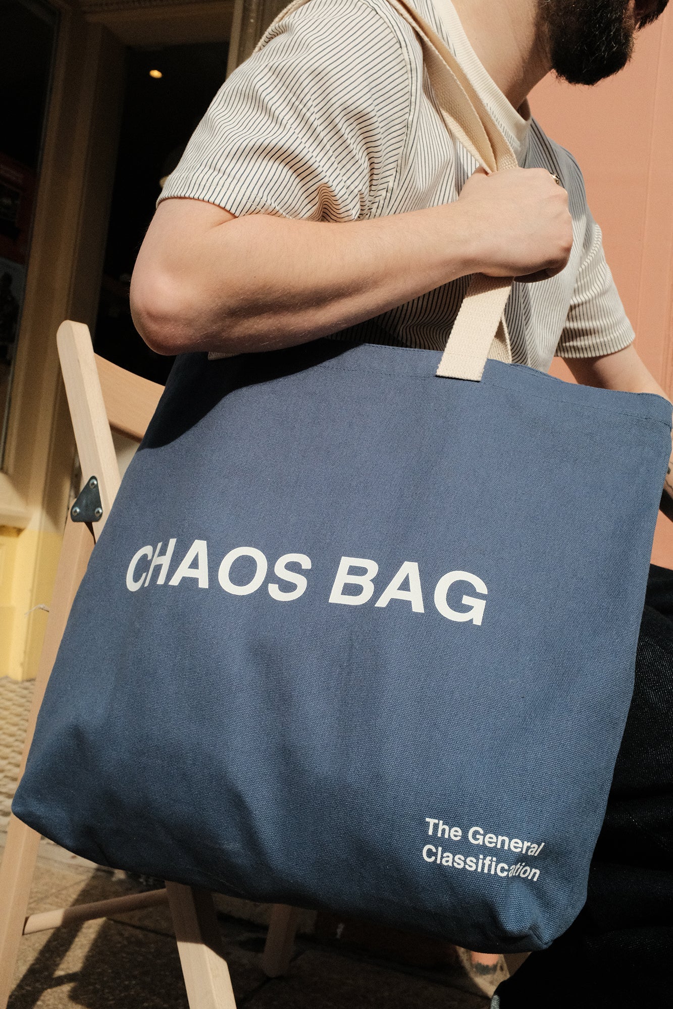 A navy blue canvas tote bag with white text reading 'CHAOS BAG' and 'The General Classification' being carried on the shoulder of a person in a striped shirt.