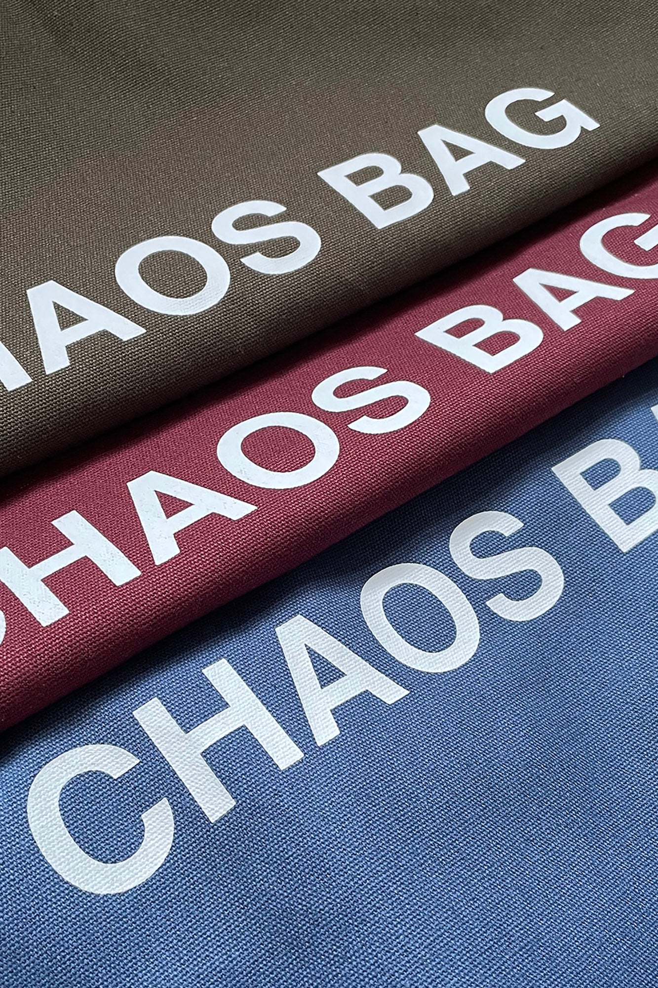 Three folded fabric bags in dark brown, burgundy, and blue, each printed with the words 'CHAOS BAG' in large white letters.
