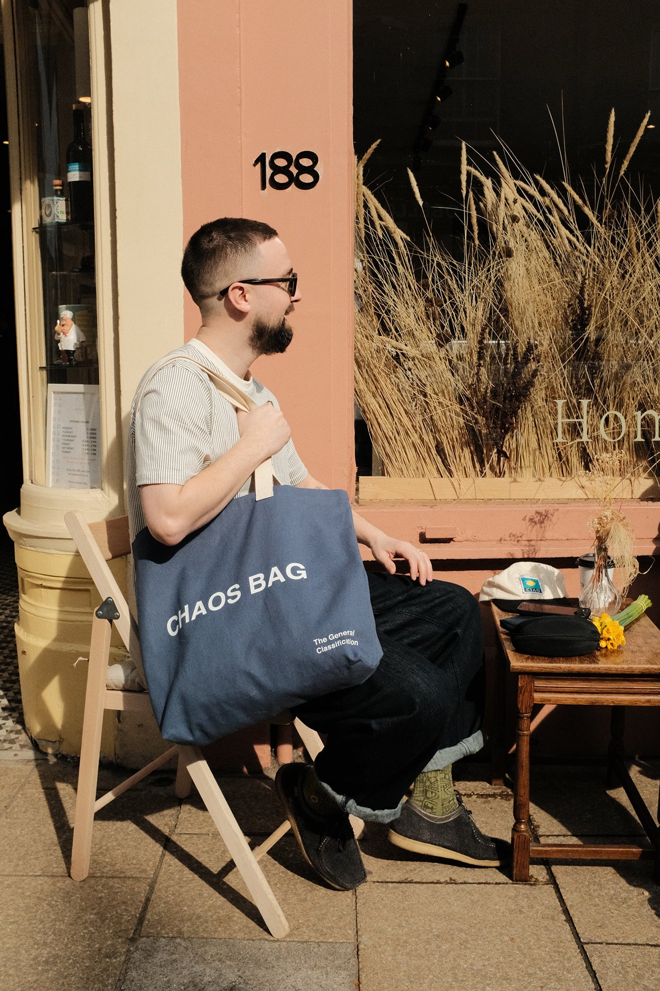 A man sat outside a cafe on a sunny day wearing sunglasses and a big latge blue tote bag with the text choas bag.