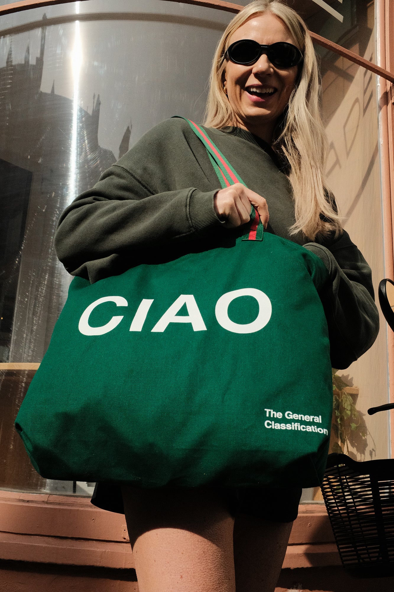 Person holding a green tote bag with 'CIAO' printed on it, standing in front of a glass door.
