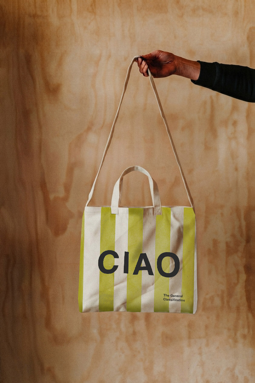A hand holding a canvas tote bag with vertical lime green and white stripes and the word 'CIAO' printed in black across the front, set against a light wood panel background.