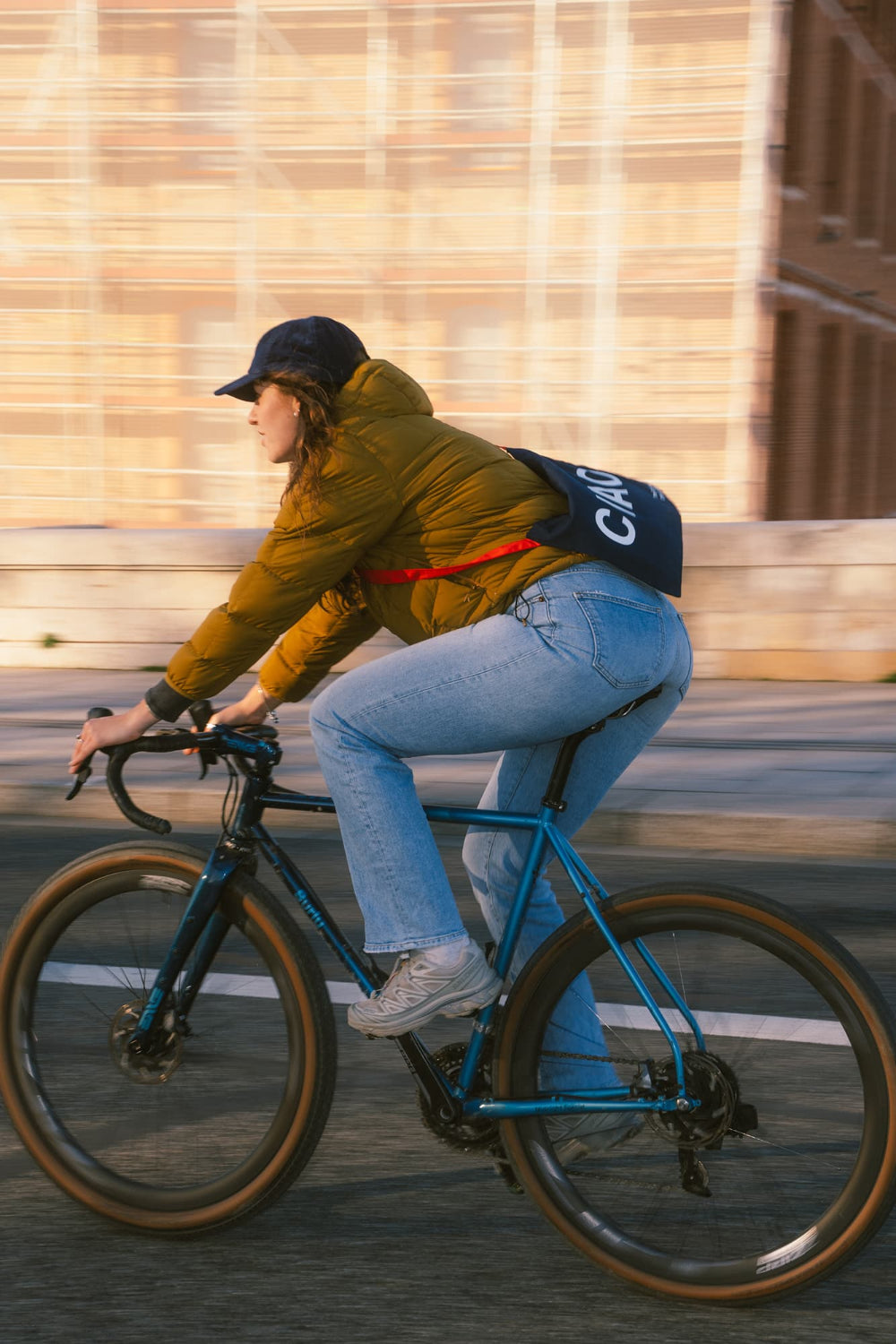 A person riding a blue bicycle outdoors, wearing a mustard yellow puffer jacket, light blue jeans, a dark cap, and a black shoulder bag with white lettering that reads CIAO against a blurred tan building background.