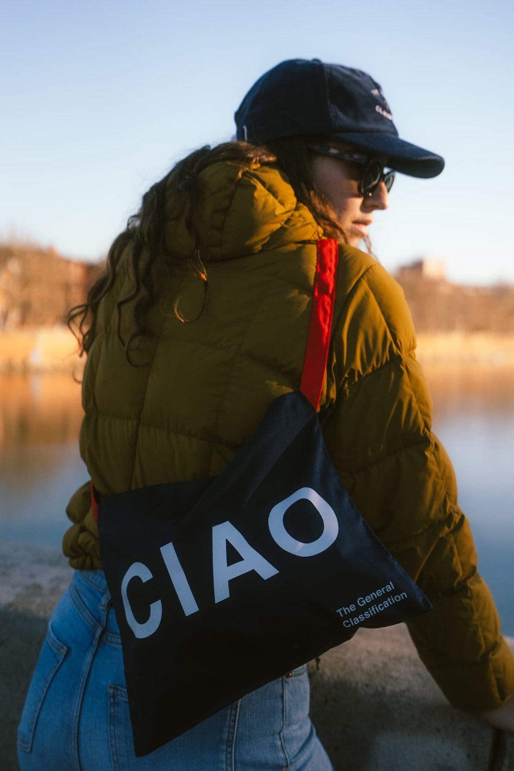 A woman wearing a black cap and sunglasses carries a black shoulder bag with the word 'CIAO' in large white letters. She is wearing an olive green puffer jacket and blue jeans, photographed outdoors near water during golden hour.