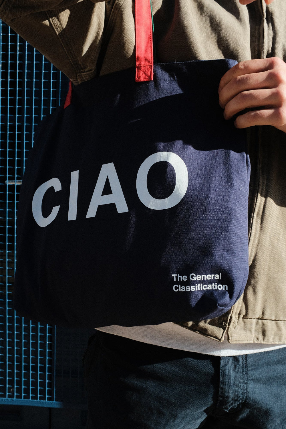A navy blue canvas tote bag with white 'CIAO' lettering and a red handle, carried by a person wearing a brown jacket and dark jeans.