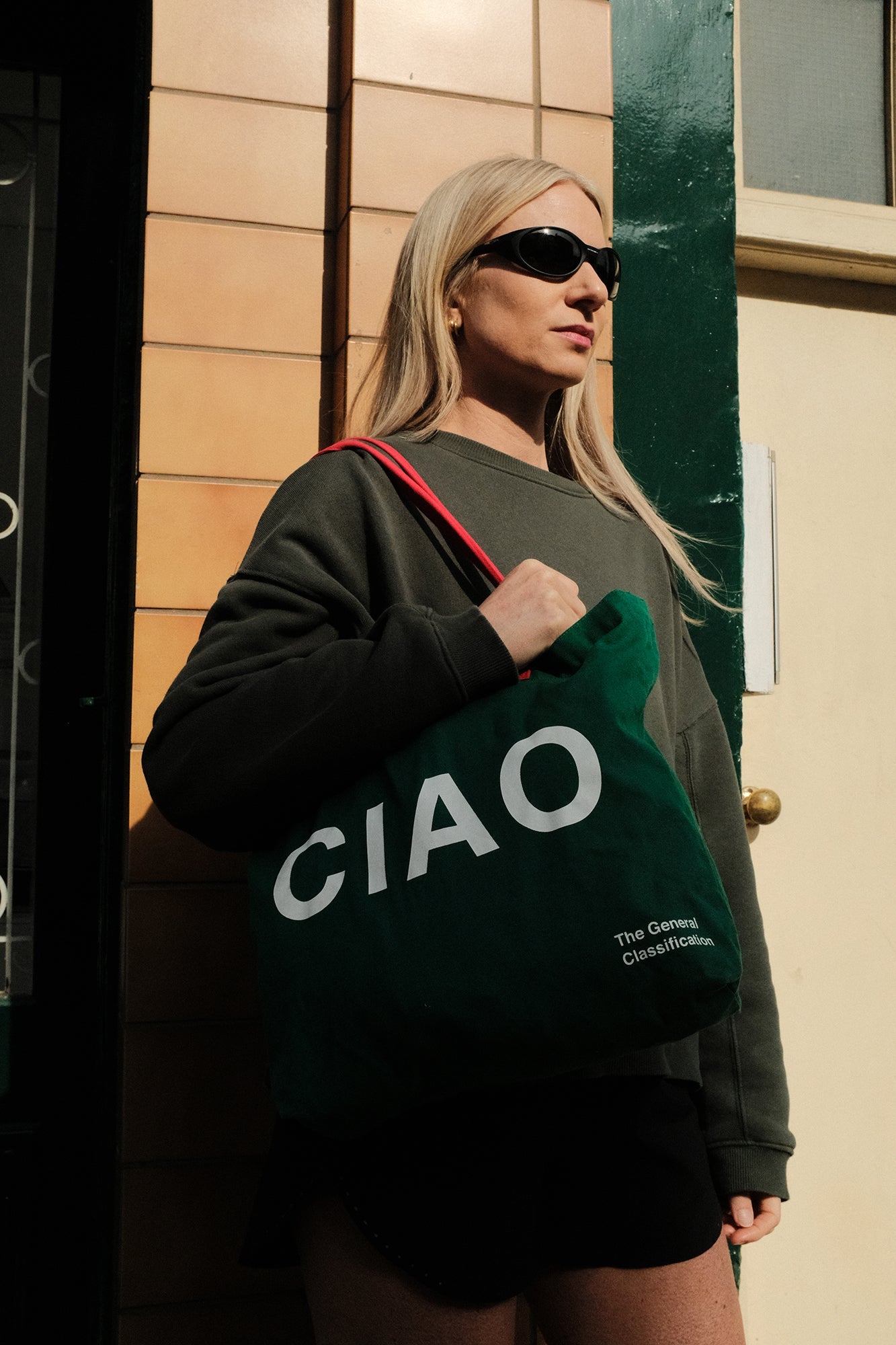 Person holding a green tote bag with 'CIAO' text, wearing sunglasses and a dark outfit.