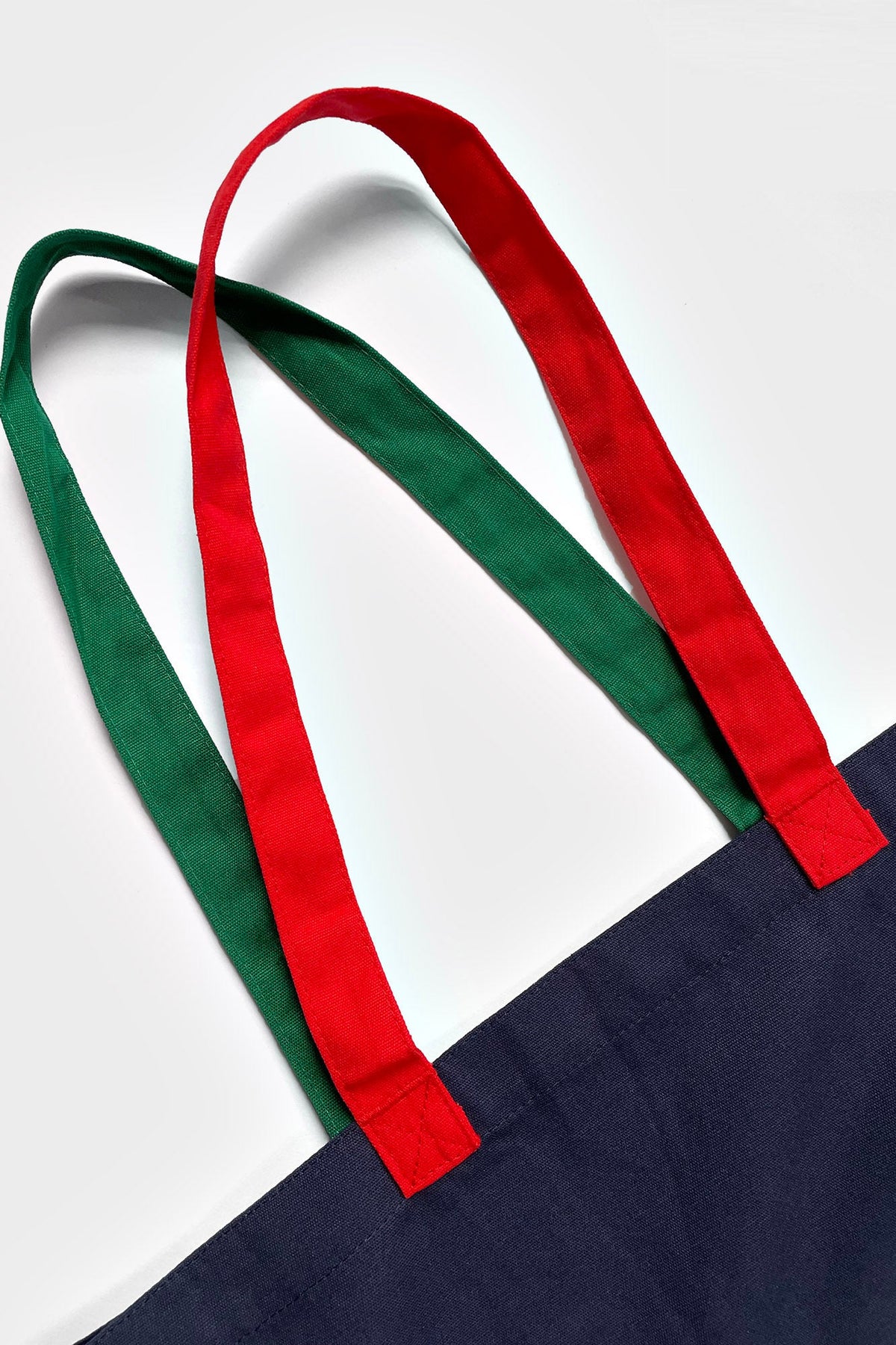 A close-up of a tote bag with a dark navy body and dual shoulder straps in green and red, shown against a clean white background.
