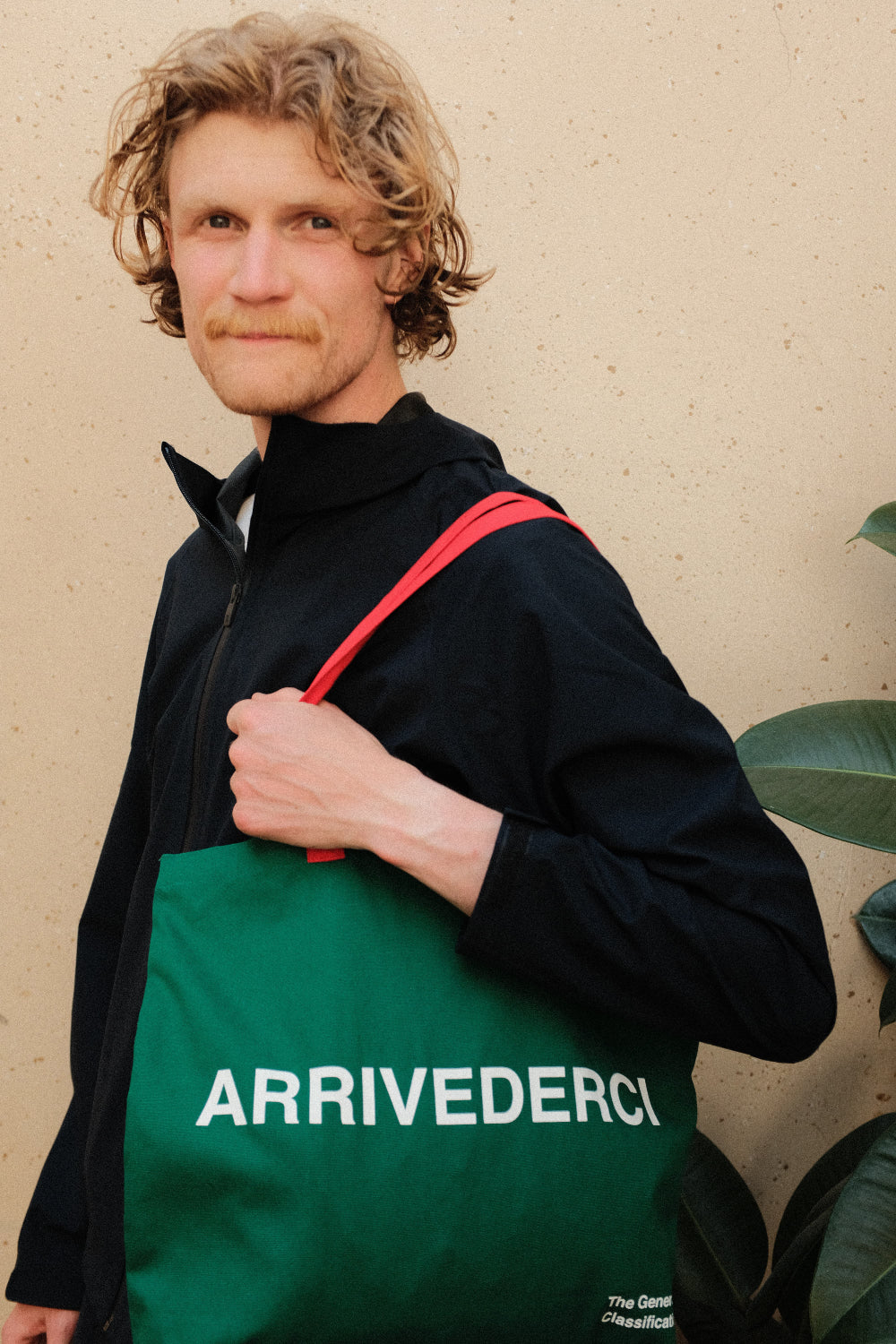 Person holding a green bag with 'ARRIVEDERCI' text against a beige wall.