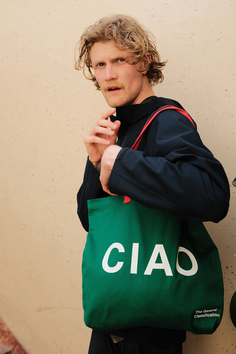 Man holding a green tote bag with 'CIAO' printed on it against a beige wall.
