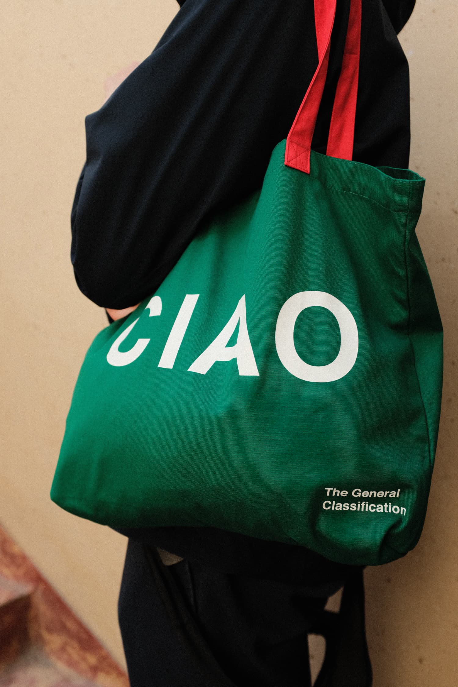 A green canvas tote bag with red shoulder straps, featuring the word 'CIAO' in large white letters and 'The General Classification' printed in white near the bottom right. The bag is being worn by a person wearing a black hoodie and black pants against a beige wall.