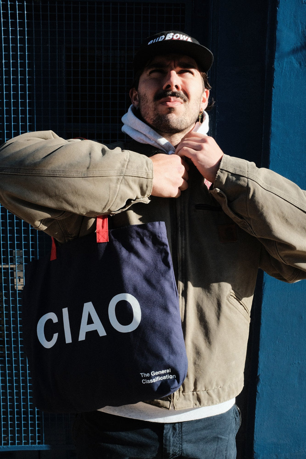 A man wearing a beige jacket and a black cap holds up a navy blue tote bag with the word 'CIAO' printed in large white letters. The background features a dark blue wire mesh screen and a solid blue wall.