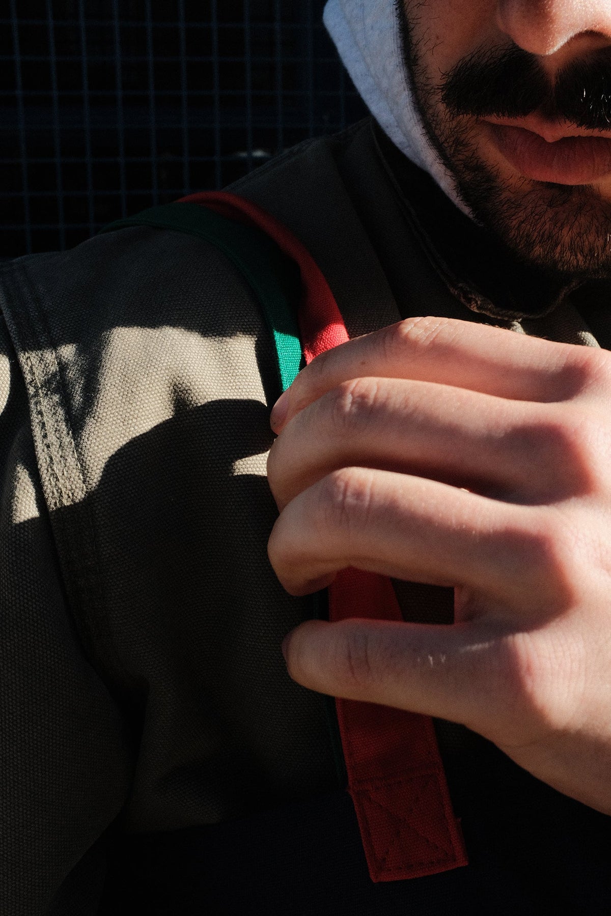 Close-up of a person wearing a dark olive green jacket with a red and green shoulder strap, holding the strap with one hand.