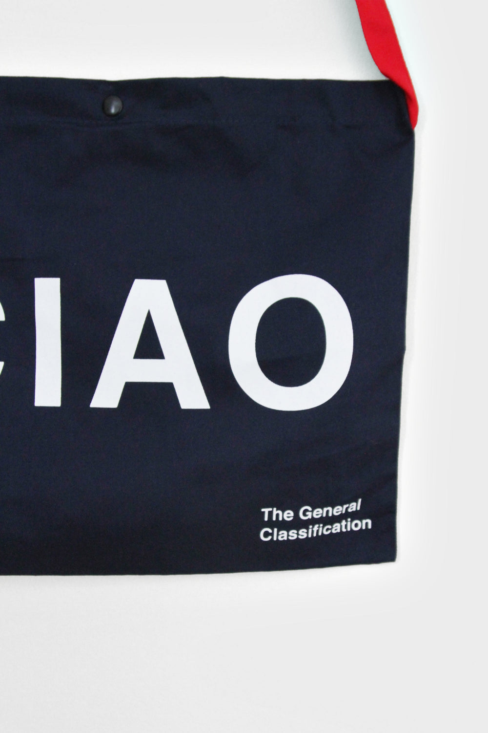 A close-up of a dark navy tote bag with bold white text reading 'CIAO' and smaller text 'The General Classification' near the bottom right. A red strap is visible at the top edge against a light gray background.