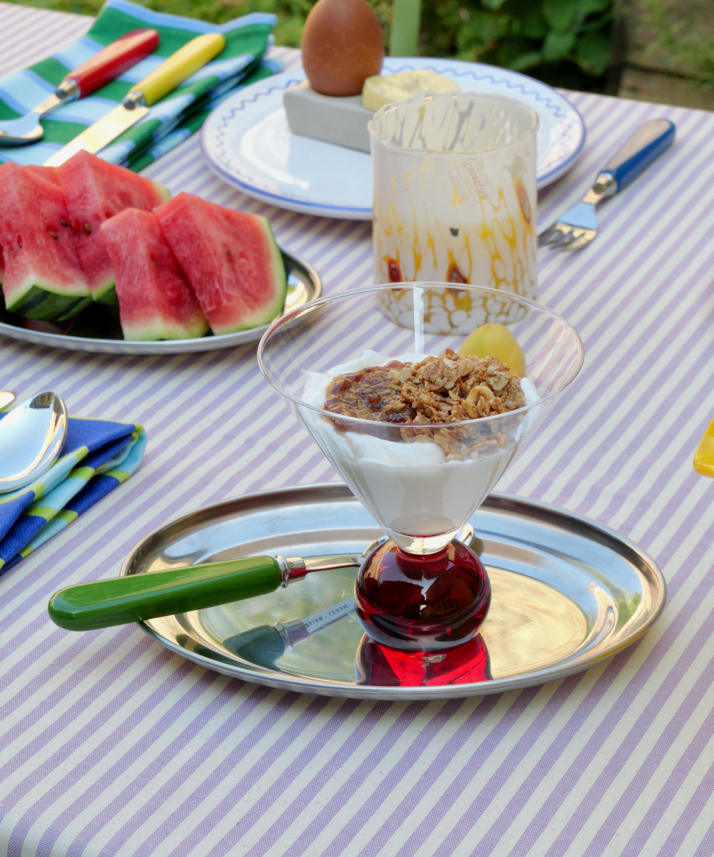 Dessert with granola and yogurt on a striped tablecloth with watermelon slices and utensils.