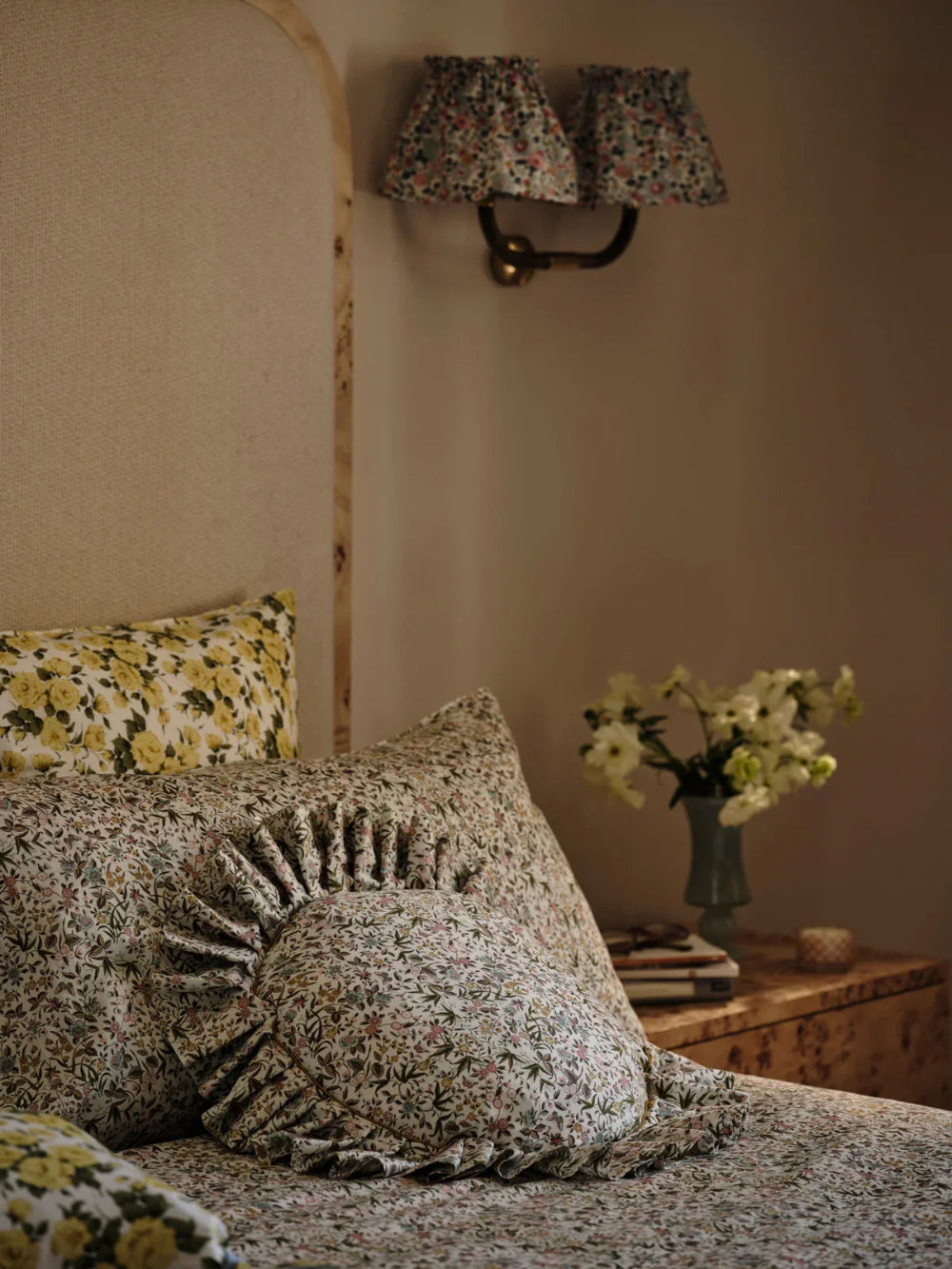Floral patterned pillows on a bed with a wall lamp and vase in the background.