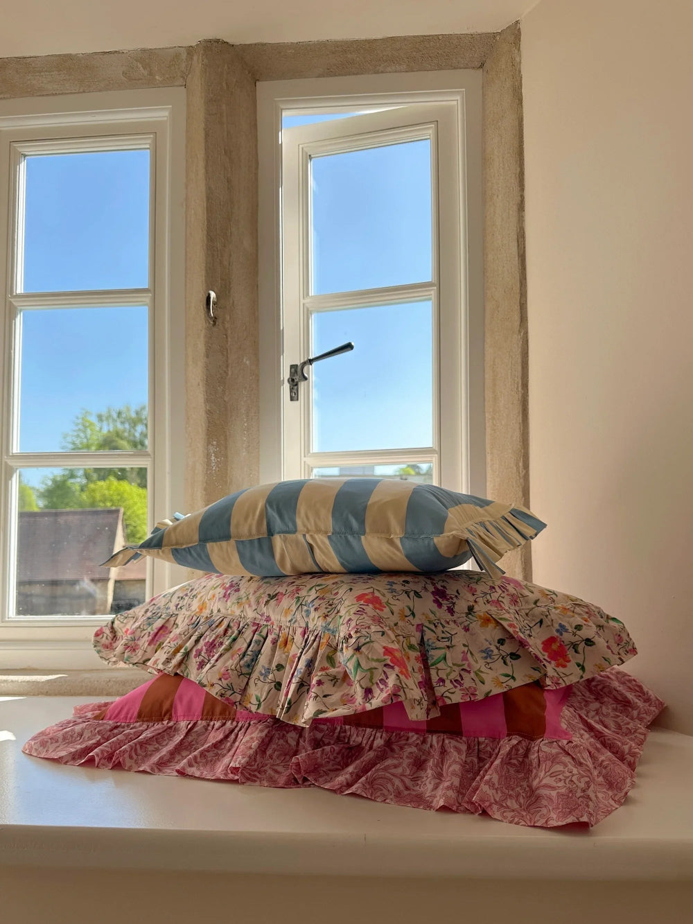 Stack of colourful pillows on a windowsill with a view of trees outside.