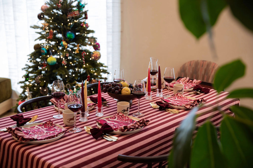 A dining table set for a holiday feast with a red and white striped tablecloth, plates of sliced cured meat, red wine glasses, and candles, with a decorated Christmas tree in the background.