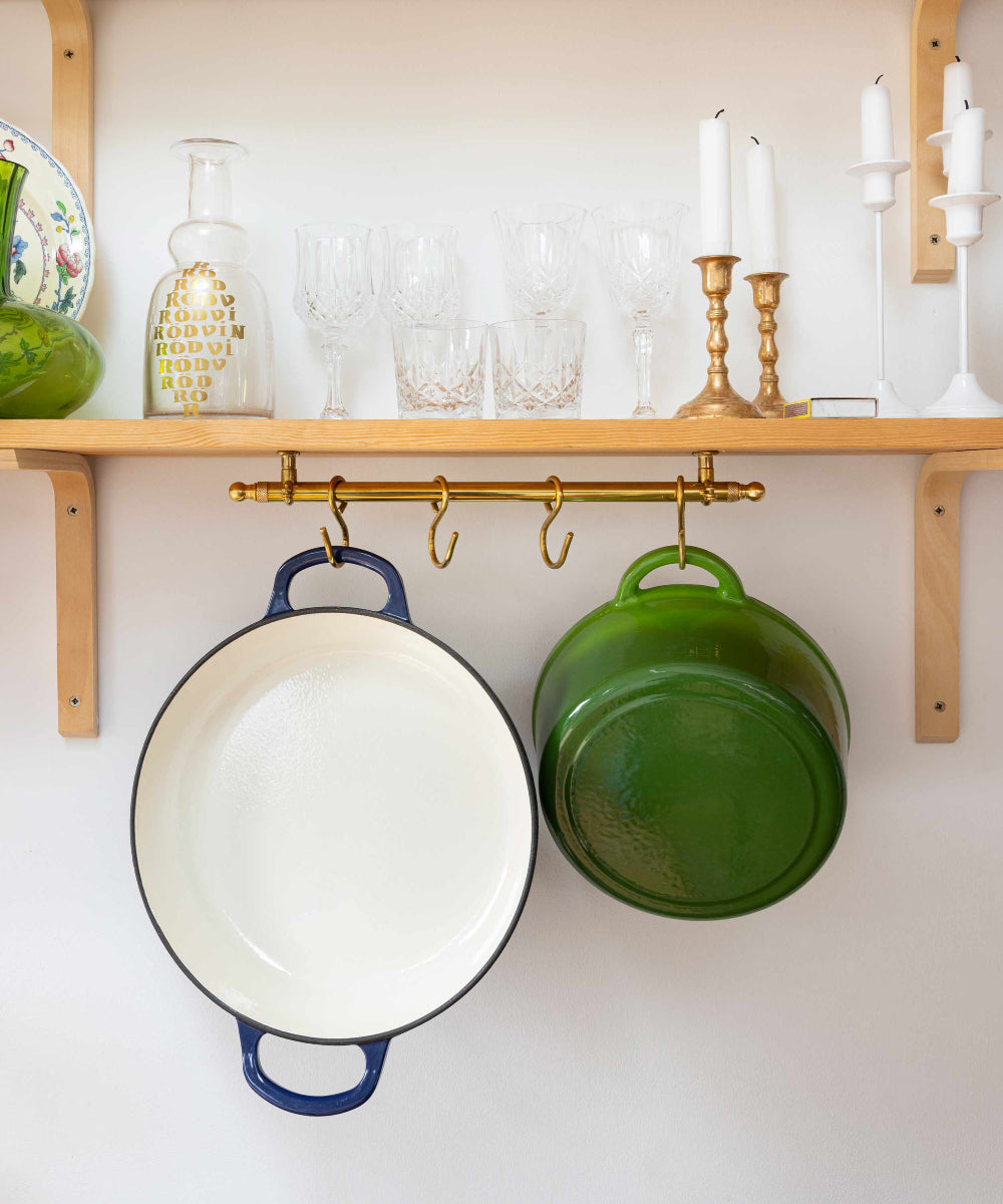 A brass wall-mounted pot rack with three hooks hanging cookware: a large white enameled cast iron pot with a blue handle, a green enameled pot upside down, and a smaller green pot hanging on the right. A light wood shelf above holds glassware, candles, and decorative bottles against a white wall.