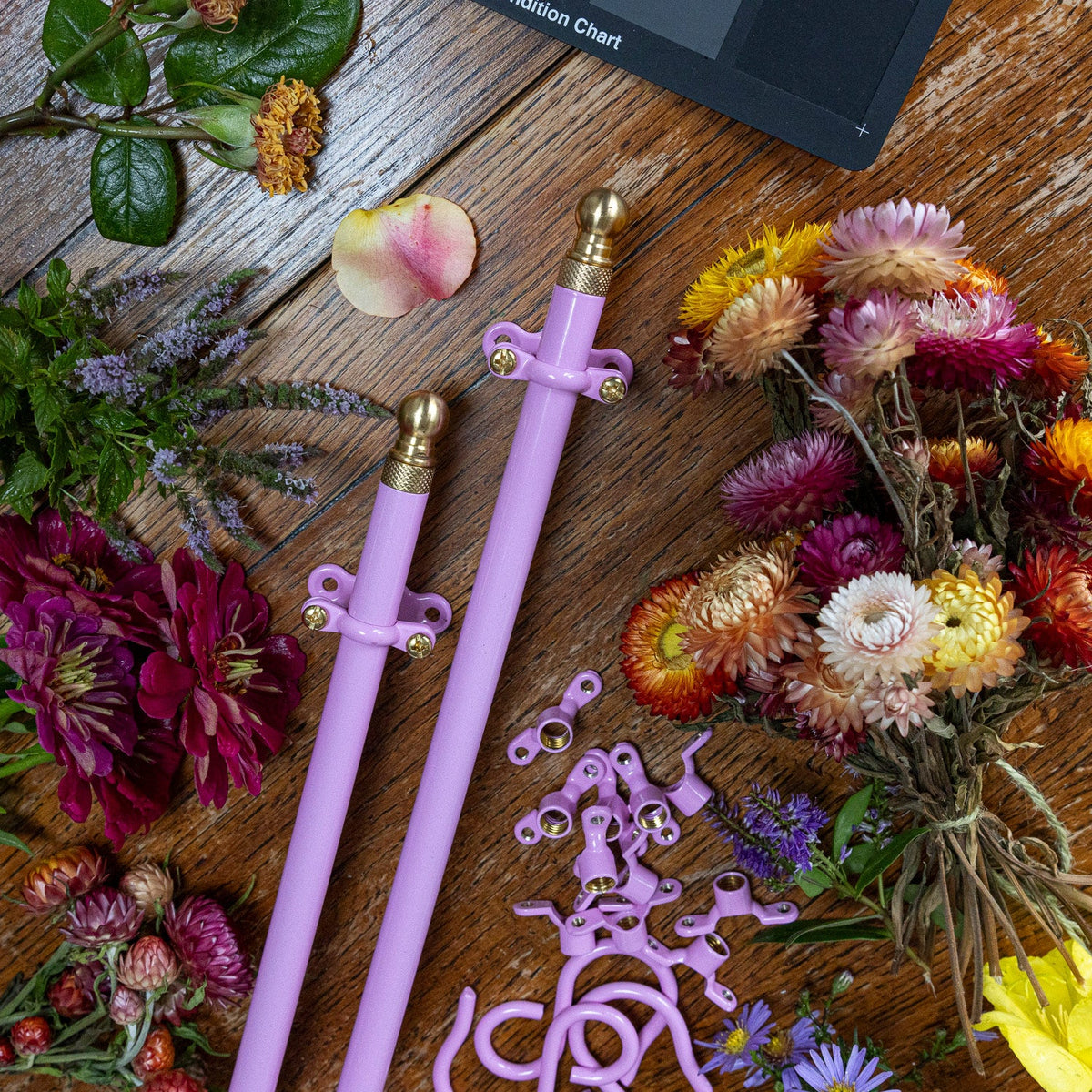 A pair of kitchen hanging rails in pink with gold-toned metal tips and matching small accessory pieces arranged on a rustic wooden surface, surrounded by colourful flowers and greenery. A partial black chart or box is visible at the top edge.