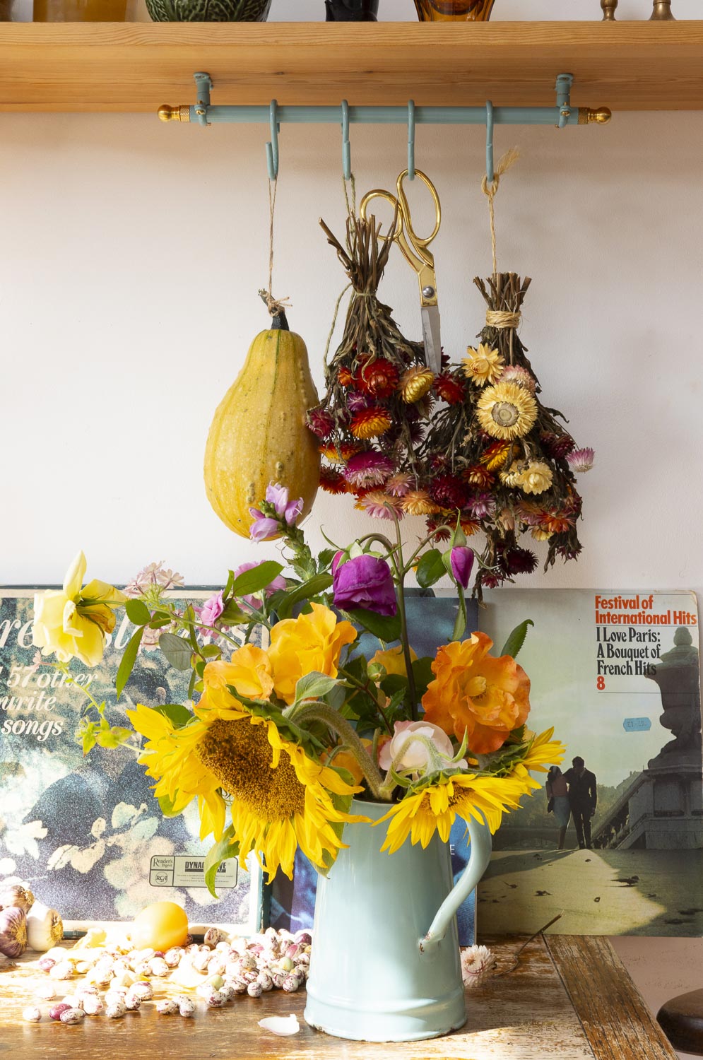A light blue metal pitcher filled with bright yellow sunflowers, orange flowers, and pink roses on a rustic wooden table. In the background, dried flowers and a yellow squash hang from a teal rack on a wooden shelf.