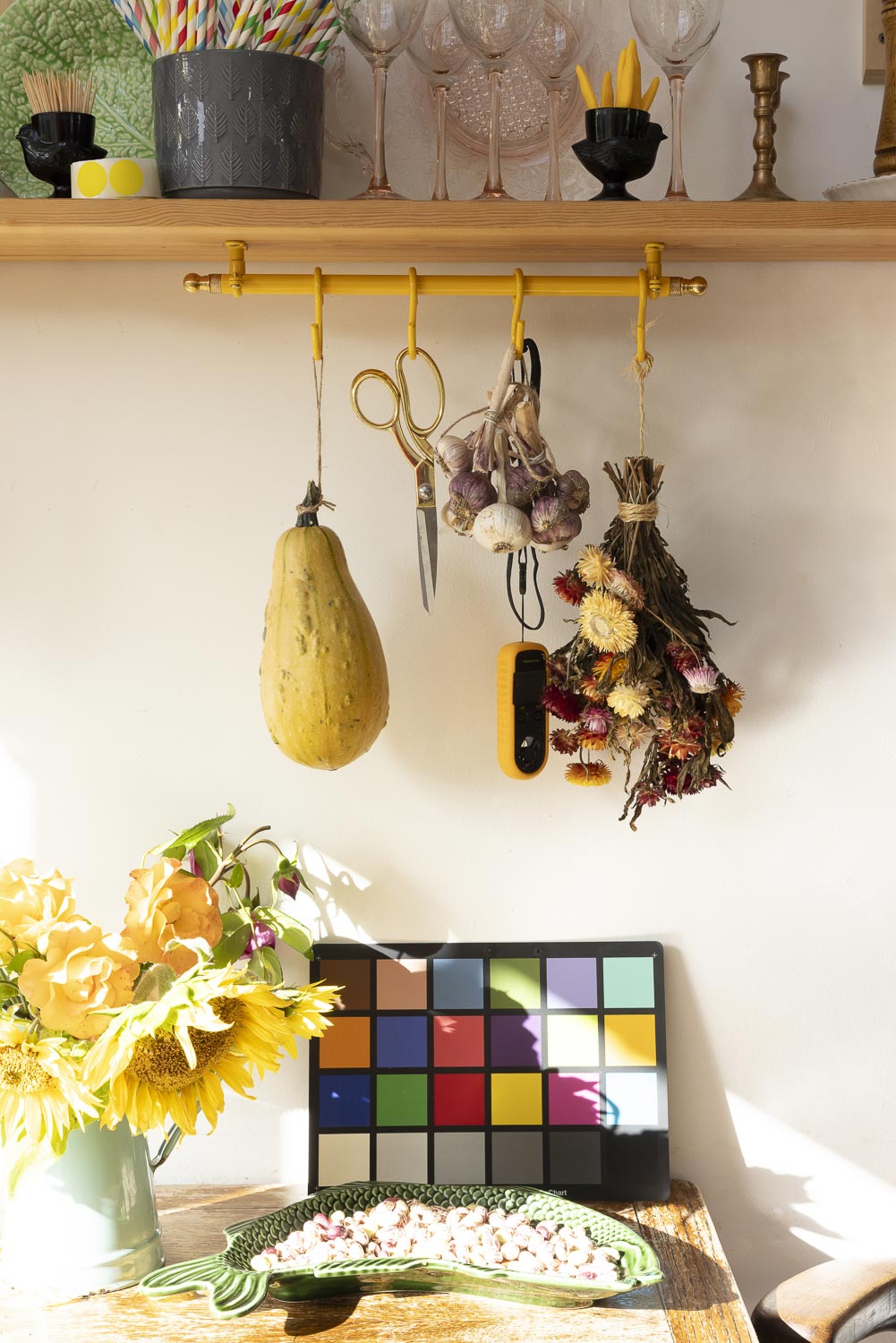 A styled interior scene with a wooden shelf holding glassware and decorative items, a yellow wall-mounted rod with hanging kitchen tools and dried herbs, a yellow squash hanging on the left, a color card board, and a green bowl on a table with yellow flowers in the foreground.