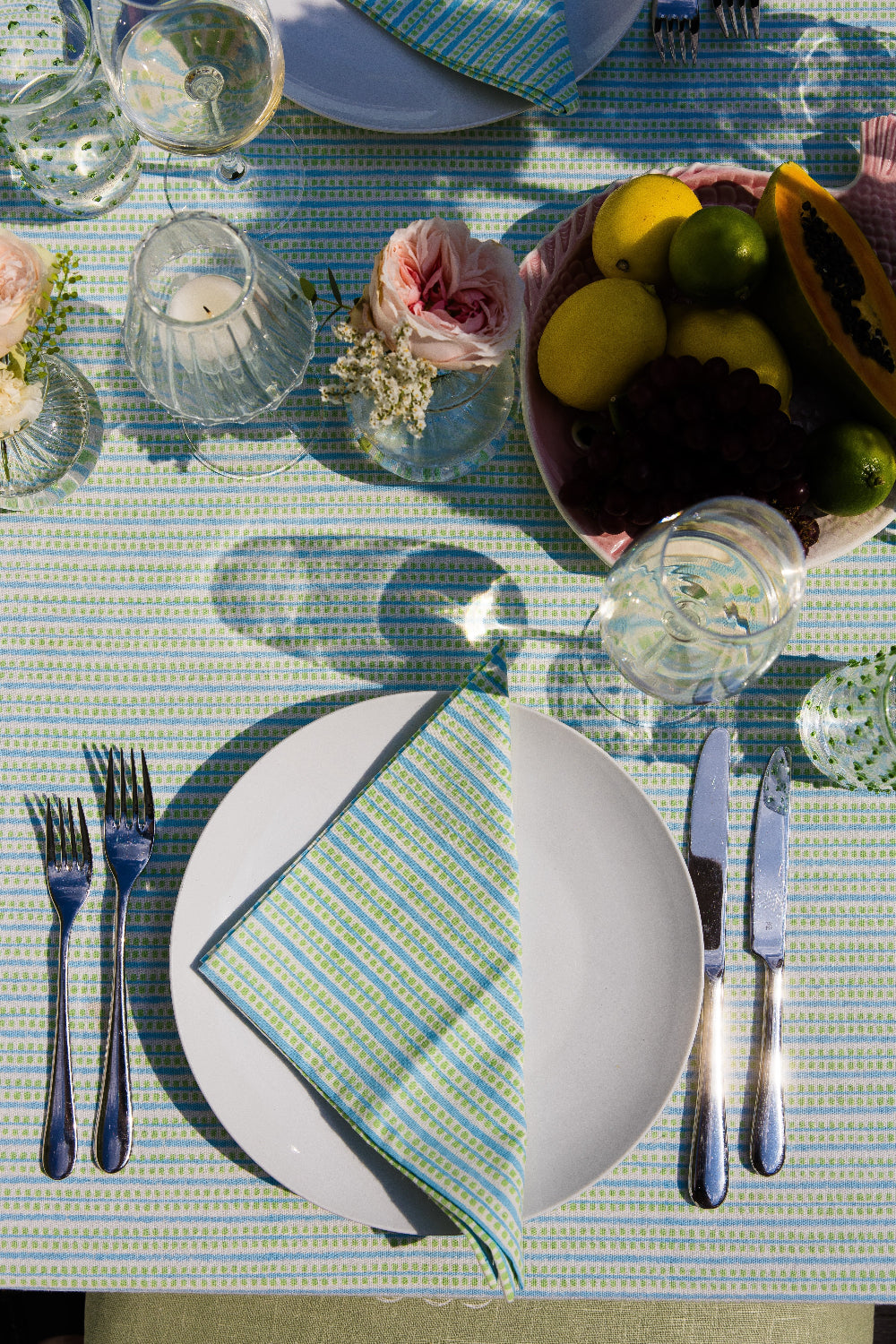 Dining table setting with plates, cutlery, and a bowl of fruit on a striped tablecloth.