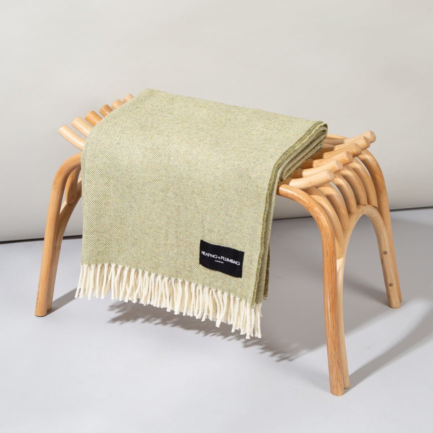 Green blanket with a brand label draped over a wooden stool on a light grey background