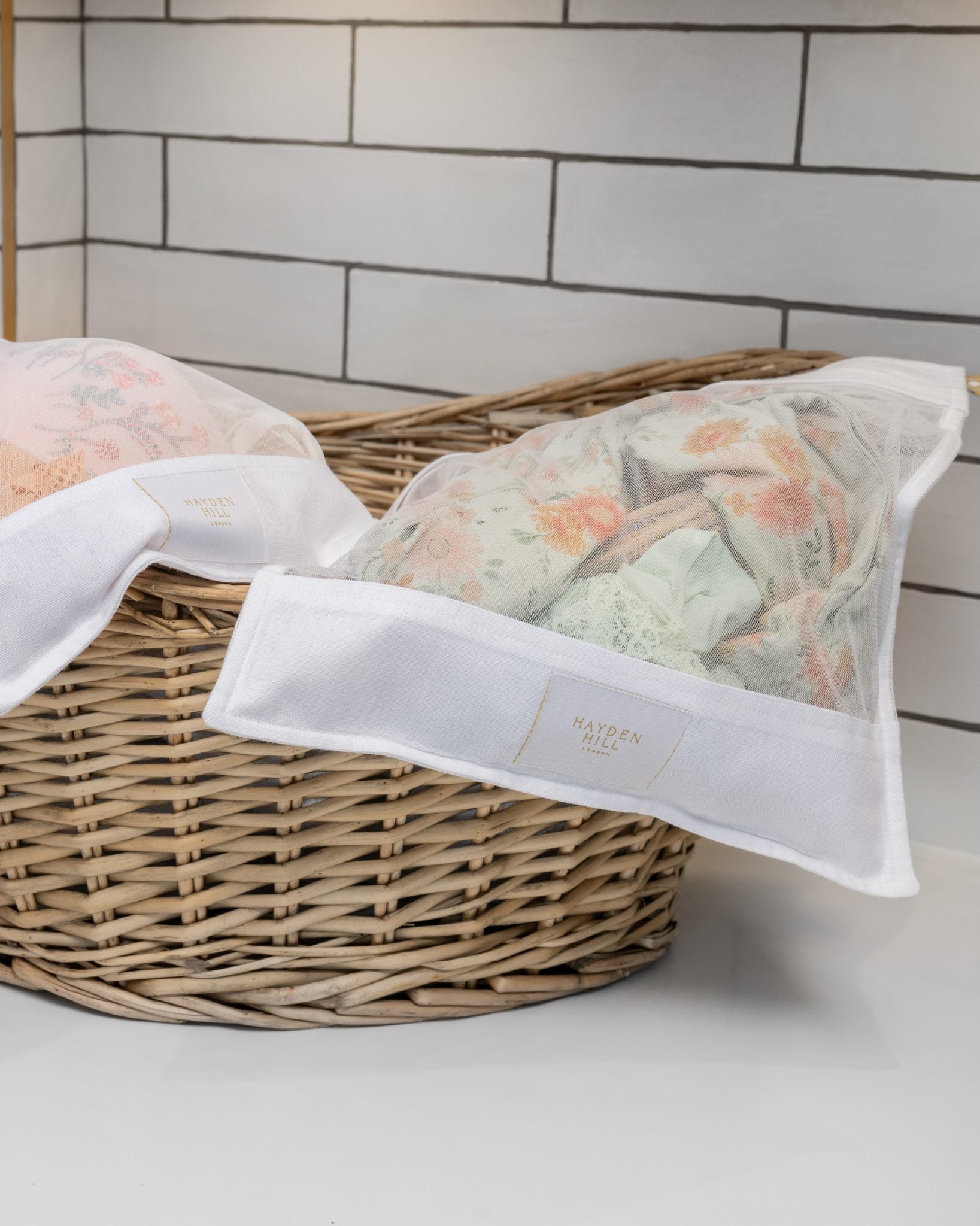 Two oragnic cotton laundry wash bags filled with patterned towels, resting in a woven wicker basket against a white subway tile background.