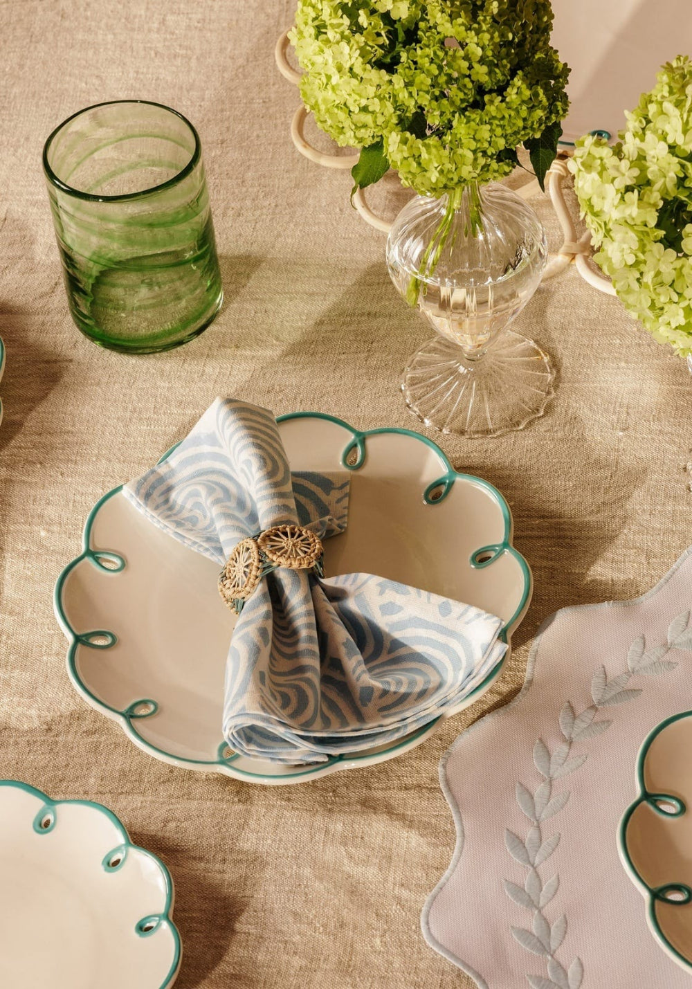 Decorative table setting with a scalloped edge plate, napkin, and green glassware on a beige tablecloth.