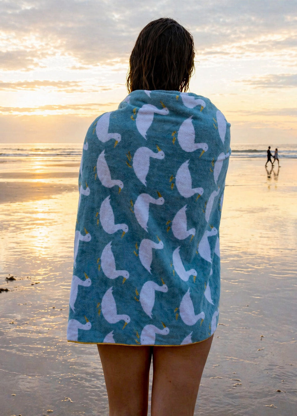 Person wrapped in a towel with a duck pattern on a beach at sunset