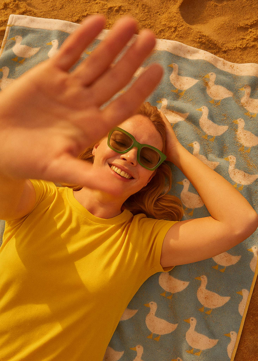 Person wearing sunglasses and a yellow shirt, lying on a towel with duck pattern at the beach.
