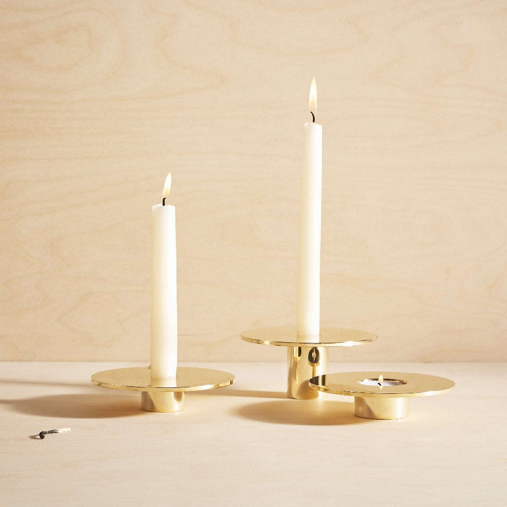 Three gold brass candle holders on a light wood surface against a matching wood background, with two white taper candles lit and one small match lying nearby.