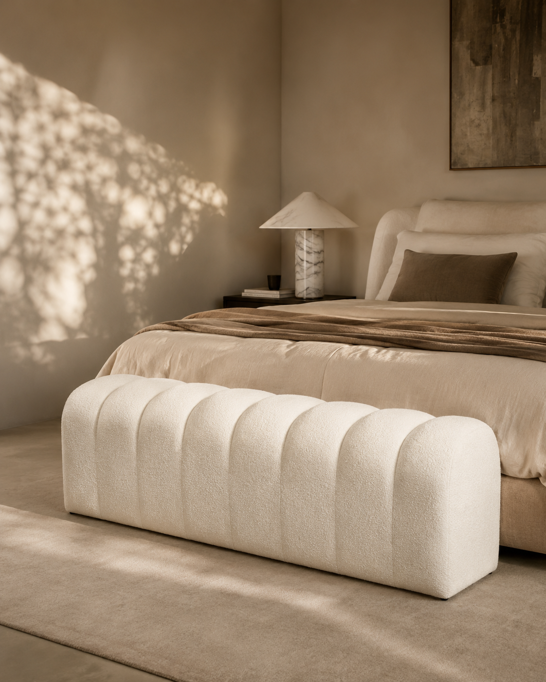 A modern bedroom scene featuring a tufted, barrel-shaped upholstered bench in a warm off-white fabric at the foot of a bed. The bed is dressed in neutral beige and brown linens, with a matching lamp on a side table and dappled sunlight casting shadows on the wall.