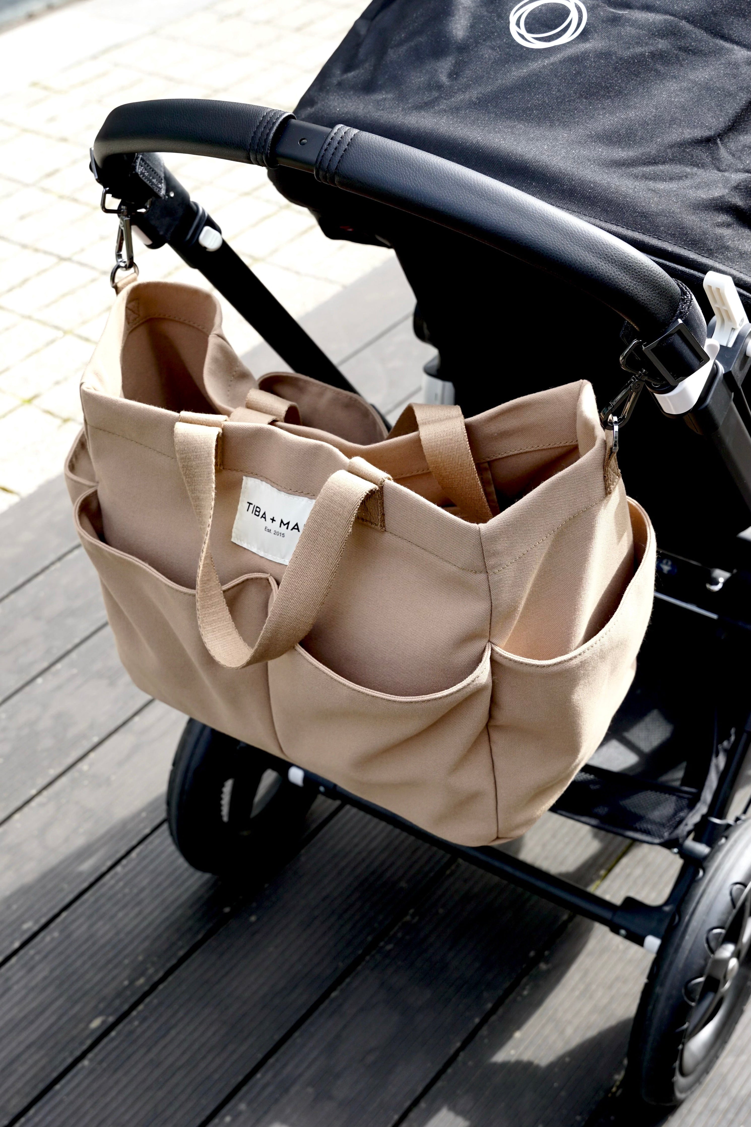 Beige Tote bag attached to a black stroller on a wooden floor.